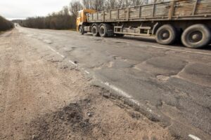 A truck towing a trailer navigates a treacherous road marred by crumbling asphalt, littered with dangerous potholes and pits, posing a hazard to drivers.